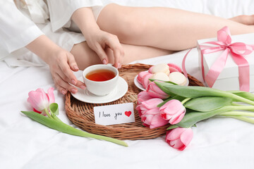 Tasty breakfast served in bed. Woman with tea, macarons, gift box, flowers and I Love You card at home, closeup