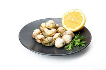 A black plate holds fresh cockles, half a lemon, and a sprig of parsley against a white background, ready for culinary use