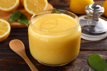 Delicious lemon curd in glass jar, fresh citrus fruits and spoon on wooden table, closeup