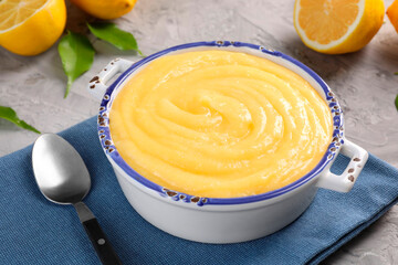 Delicious lemon curd in bowl, fresh citrus fruit, green leaves and spoon on grey table, closeup