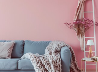 A beige and gray knitted blanket lies on a blue sofa against a pink wall in a living room interior with a white ladder, table lamp and decorative elements
