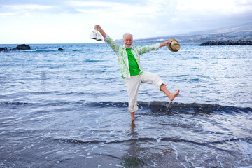 Happy cheerful elderly man barefoot on the water's edge still fully dressed while jokingly lifting...