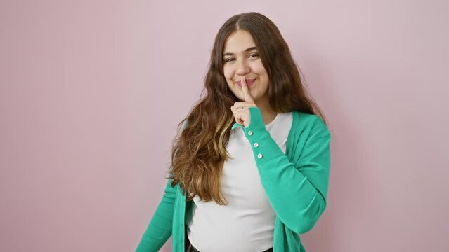 Shush! beautiful young hispanic woman confidently stands in silence, finger on lips gesturing a secret quiet. over a calm, isolated pink background. relax, stop talking, striking a silent pose.