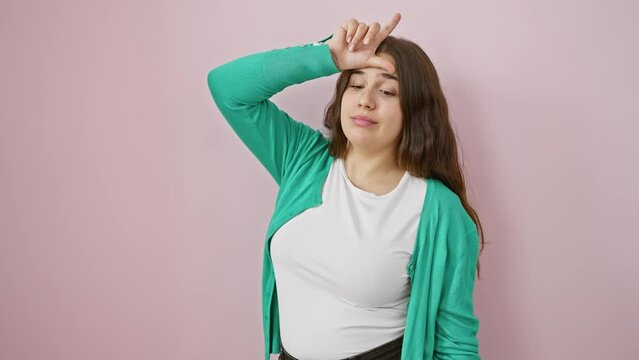 Young beautiful hispanic woman standing making fun of people with fingers on forehead doing loser gesture mocking and insulting. over isolated pink background