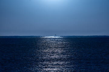 Clear, bluish night sky illuminated by moonlight with its rays of light beautifully reflected in the water of the Mediterranean Sea and a backlit fishing boat silhouette on the horizon line.