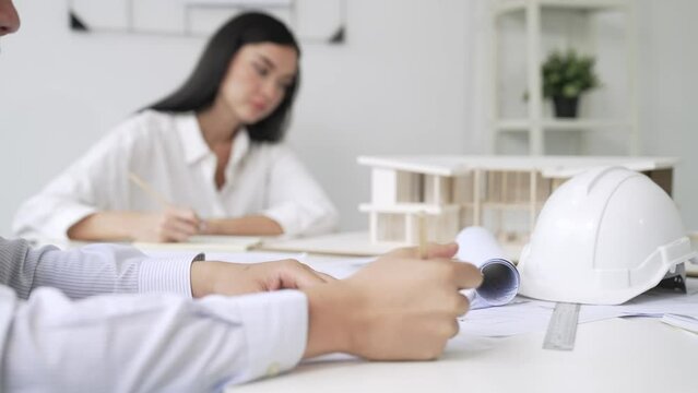Professional architect engineer drawing blueprint and working together with young beautiful caucasian coworker on meeting table with safety helmet, house model, blueprint scatter around. Immaculate.