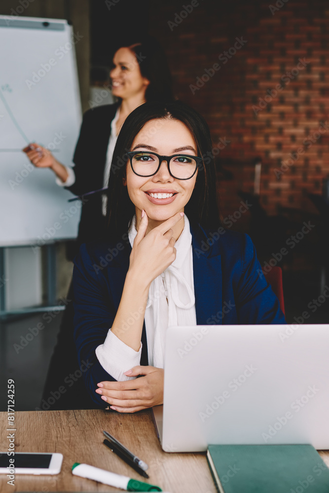 Poster portrait of cheerful female manager in spectacles for vision correction sitting at desktop with lapt
