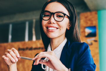 Half length portrait of cheerful caucasian businesswoman holding pen in hands and looking at camera with cute smile on face, young successful female coworker enjoying management in company office - Powered by Adobe