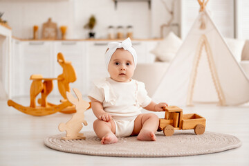 A cute baby girl of six months is playing sitting on the floor with wooden toys in white clothes in a bright children's room at home, a happy healthy child with a typewriter