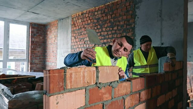 Construction Worker Building a Brick Wall, A focused construction worker in a reflective vest meticulously lays bricks on a partially built wall inside a building under renovation.