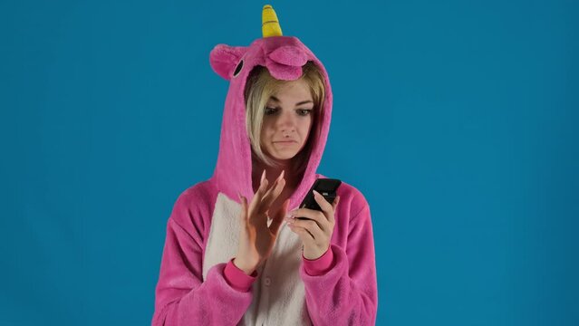 Female teenager in unicorn pajamas examines old smartphone on blue background. Confused girl tries to make call unsuccessfully and throws phone