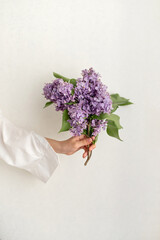 Minimalist aesthetic floral business branding design, lilac flowers bouquet in woman hand on light creamy wall background with soft natural light and shadows, spring gift or greeting card
