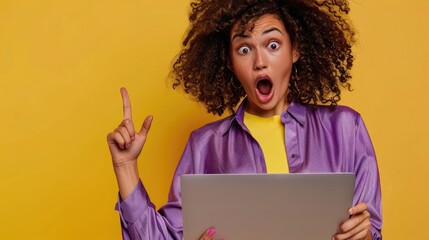 Young woman holding a laptop and pointing her finger to the side, against a yellow background, wearing a purple blouse, with curly hair, a surprised expression, open mouth, looking at the camera