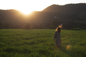 Woman with long coat holding long hair enjoying sunset on the green meadow