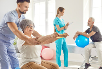 Nurse helping senior smiling woman to do sport stretching exercises with rubber band sitting on fit ball in clinic. Physiotherapist or health care worker helping elderly mature people in rehab.