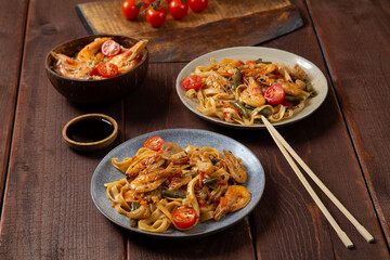 Rice with shrimp and udon in yaki soba sauce on a wooden table next to cherry tomatoes, soy sauce and chopsticks. Horizontal