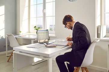 Accountant in a modern office workplace. Man in a suit sitting at his working desk, studying information in a thick paper binder and using business sheets on his laptop computer