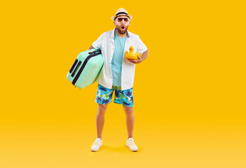 Surprised summer tourist. Full length bearded man in white blue holiday wear, sun hat and sunglasses holding toy and green travel bag standing with amazed face expression on yellow orange background