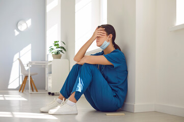 Exhausted stressed female nurse or doctor wearing blue face mask and uniform sitting with closed...