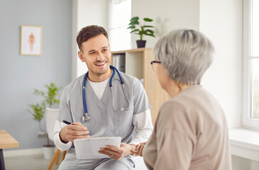 Friendly doctor gives informative medical consultation to elderly woman during clinic appointment. Smiling doctor with clipboard tells mature patient about treatment and explains examination results.