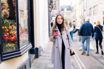 Young smiling woman in trench coat enjoying hot coffee drink in reusable cup while walking on the...