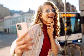 Beautiful woman in good mood looks into camera and takes selfie in the street. Blogging,...