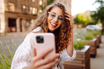 Beautiful woman in good mood looks into camera and takes selfie in the street. Blogging,...