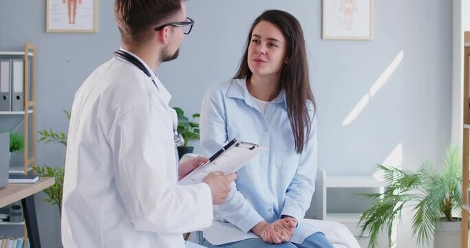 Portrait of a male doctor talking with young female patient sitting on couch in clinic. Physician giving consultation a woman during medical examination in office. Medicine concept. 4k video.