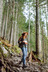 Naklejka premium Young female traveler with backpack standing in forest with tall coniferous trees, enjoying nature. Weekend, tourism. 