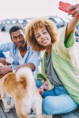 Cheerful african american hipster guys together with dog making selfie photo on front camera of modern smartphone.Positive young man and woman with curly hair shooting video on telephone for blog