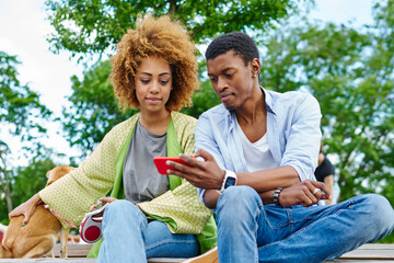 Pensive african american young man and woman dressed in casual wear spending free time together with dog sitting on wooden bench and watching video online on modern smartphone using 4g internet