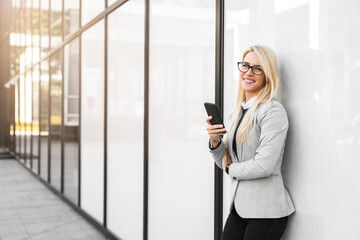 Attractive young blonde female bank manager using mobile phone while leaning on white modern wall...