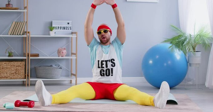 Funny and nerdy man in 90s sportswear is sitting on the floor, doing stretching exercises and splits. Engaged in fitness and sport training, blending nostalgia with workout routine.