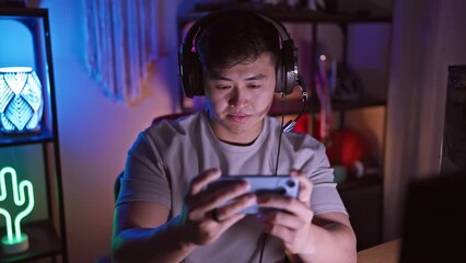 A focused asian man wearing headphones plays a mobile game in a dimly lit gaming room at night.