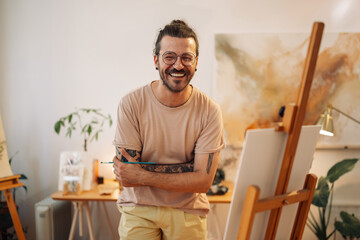 Portrait of smiling hipster artist with arms crossed standing at studio
