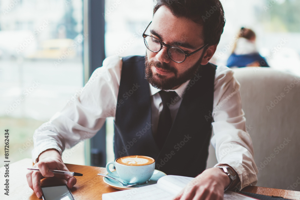Poster young hipster guy in spectacles searching interesting news on smartphone device for writing in noteb
