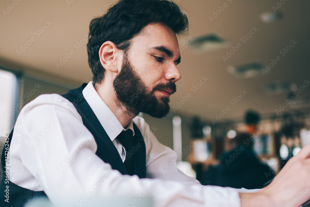 Poster serious bearded man searching information in internet web page on cellphone while sitting at table i