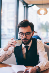 Concentrated hipster guy in eyeglasses for vision correction holding cup with hot tea during free...