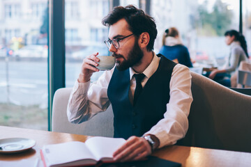 Fashionable hipster guy drinking hot coffee and looking at cafeteria window after reading...