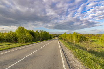 Asphalt road through a field, Evening landscape. Blue sky with clouds above the horizon. early spring in the countryside, soft sunlight on the grass.