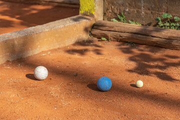 The petanque balls sit on the clay playing surface, casting shadows in the sunlight, against a backdrop of wood, logs and stone walls, suggesting a casual game of petanque.