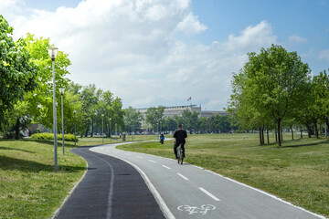  A cyclist enjoys a sunny day on a well-maintained bike path surrounded by lush greenery and trees in an urban park, with a building and flagpole visible in the distance.