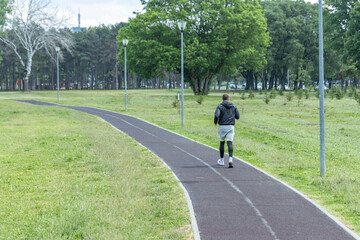 The recreational path is lined with lampposts, and in the distance, a playground and parked cars are visible. The sky is overcast, providing a calm and serene atmosphere for the jogger...