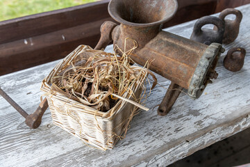 An old-fashioned manual grinder sits on a weathered wooden bench beside a basket filled with dried grass, surrounded by various metal tools, evoking a sense of rural life and traditional craftsmanship