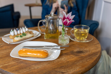 A casual café setting with a wooden table featuring an eclair on a white plate, accompanied by silver cutlery and a glass cup.