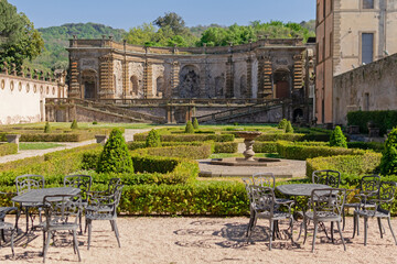A beautiful summer park with figured trees and paths in the courtyard of an ancient villa, fenced with an ancient stone wall with sculptures, there is figured garden furniture made of metal