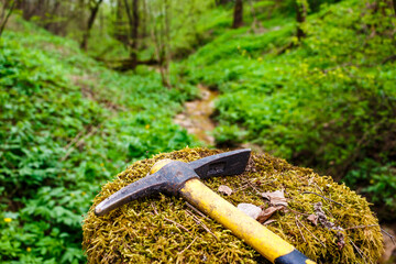 A pickaxe lying on a stump overgrown with green moss against the backdrop of a forest ravine,...