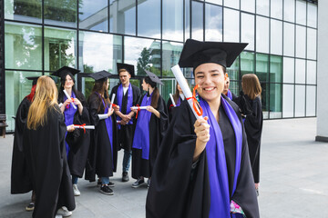 Portrait of graduate student standing in front of her friends with toga and diploma all happy.