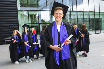 Portrait of graduate student standing in front of his friends with toga and diploma all happy.