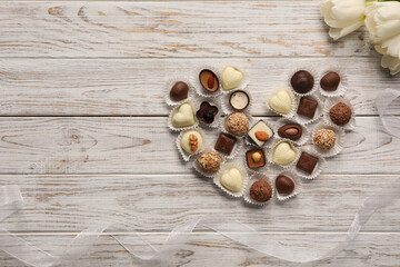 Heart made with delicious chocolate candies and beautiful tulips on white wooden table, flat lay. Space for text